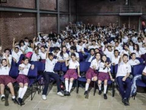Gran grupo de estudiantes sentados en medio del evento de sensibilización de seguridad Digital de CiberPaz, muchos de ellos levantando las manos y sonriendo. La mayoría viste uniforme escolar, y el ambiente es de entusiasmo colectivo.