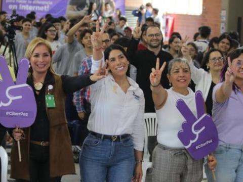 Líderes y asistentes al evento “Ciberpaz” levantan las manos haciendo el signo de la paz, rodeados de estudiantes y docentes en un ambiente de alegría y participación.