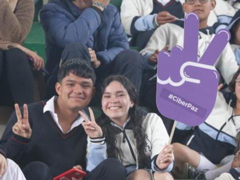 Estudiantes adolescentes sonríen mientras levantan símbolos de paz y un cartel con el mensaje #CiberPaz, sentados en las graderías del evento.