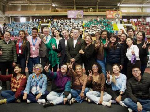 Docentes, organizadores y líderes comunitarios posan junto al escenario del evento de CiberPaz en Soacha, mientras al fondo se ve el coliseo lleno de estudiantes por colores institucionales.